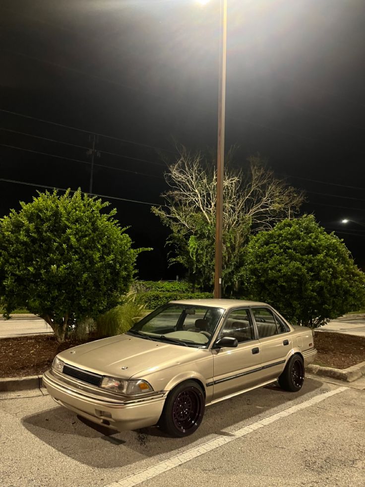 a beige car parked in a parking lot at night with the lights on and trees behind it