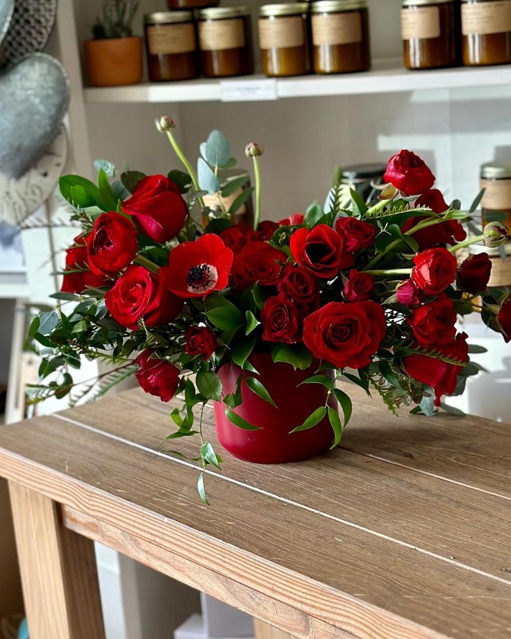 a vase filled with red roses on top of a wooden table next to shelves full of jars