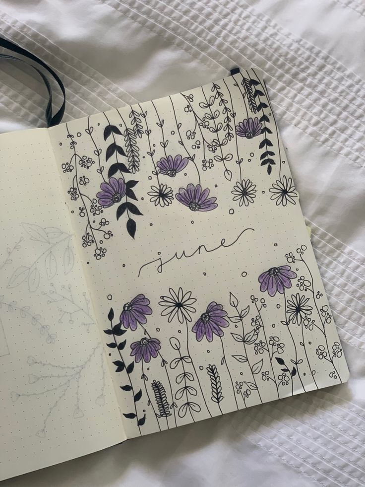 an open notebook with purple flowers on it sitting on a white bed sheet next to a pen