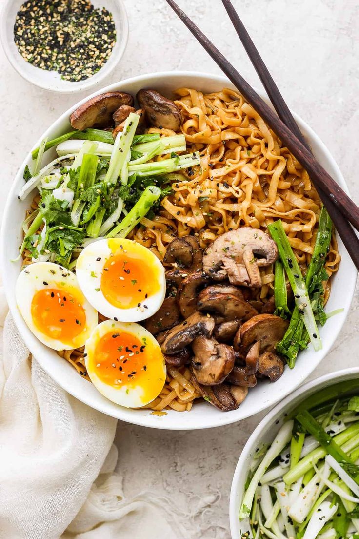 two bowls filled with noodles and vegetables next to chopsticks on a white surface