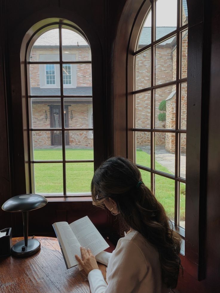 a woman sitting at a table reading a book in front of two windows with an outside view