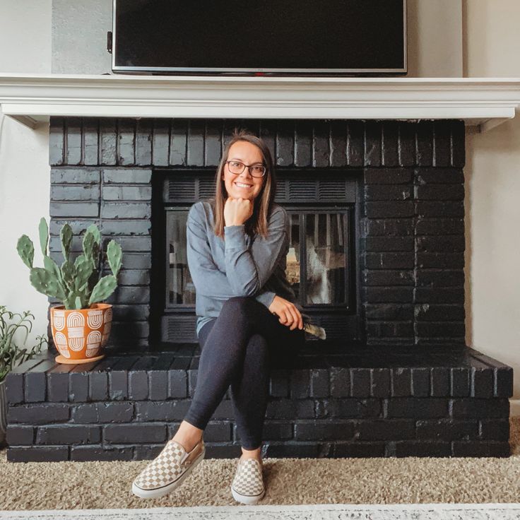 a woman sitting in front of a fireplace with her hand on her chin and looking at the camera