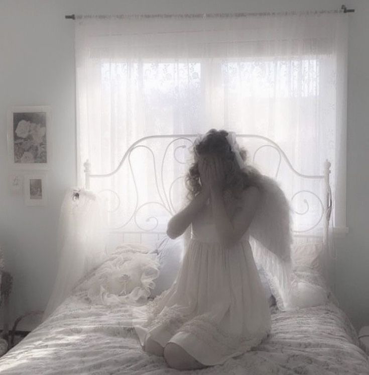 a woman sitting on top of a bed in front of a window with white curtains