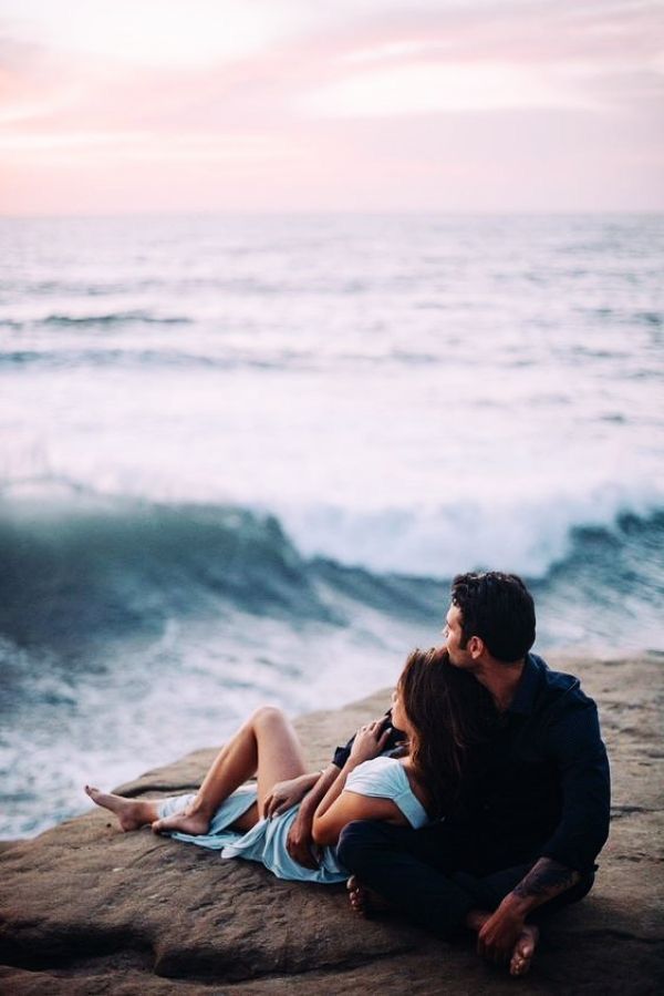 a man and woman sitting on top of a rock next to the ocean at sunset