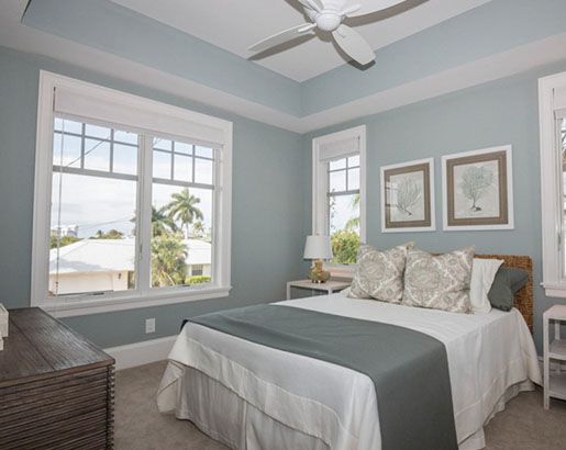 a bed sitting in a bedroom next to two windows with paintings on the wall above it