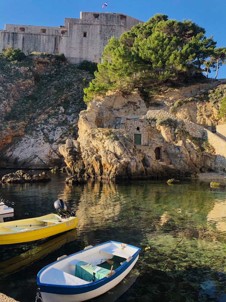 three small boats are docked in the water near a rock wall and an old castle