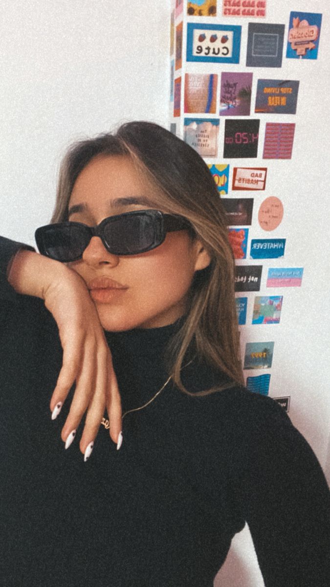 a woman in black shirt and sunglasses leaning against a wall with posters on the wall behind her