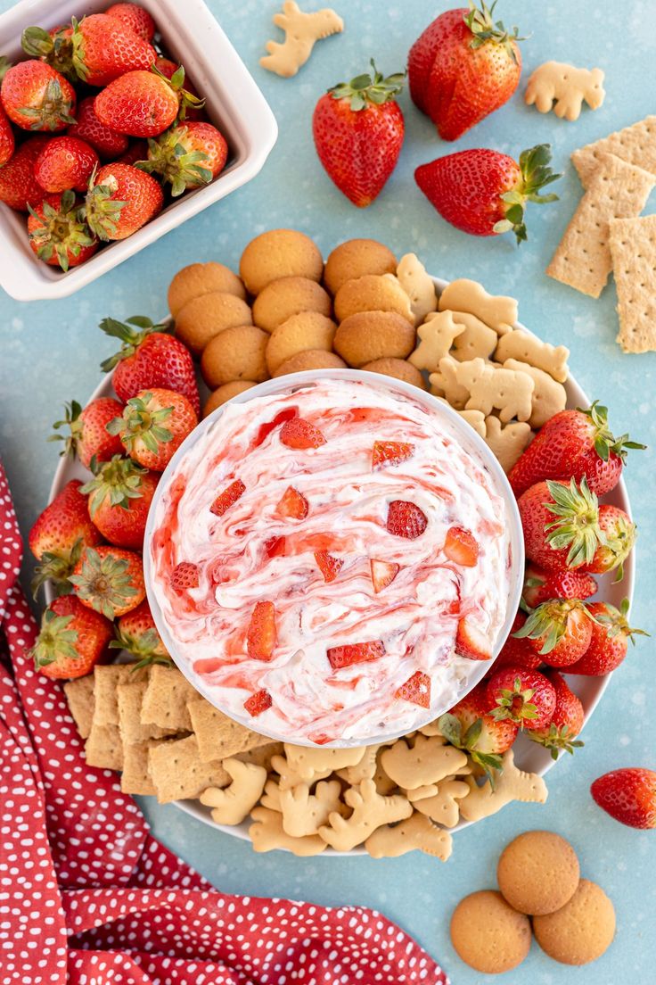 a bowl of dip surrounded by crackers and strawberries on a blue tablecloth