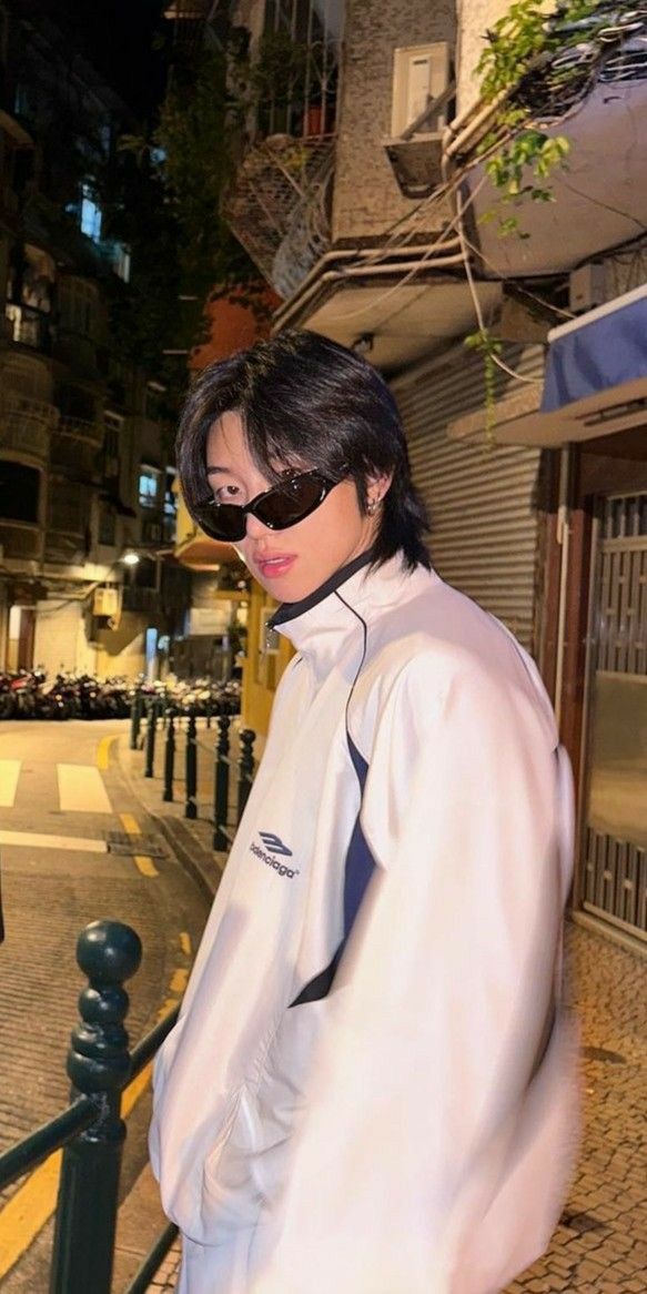 a woman standing on the side of a street next to a fence wearing sunglasses and a white jacket