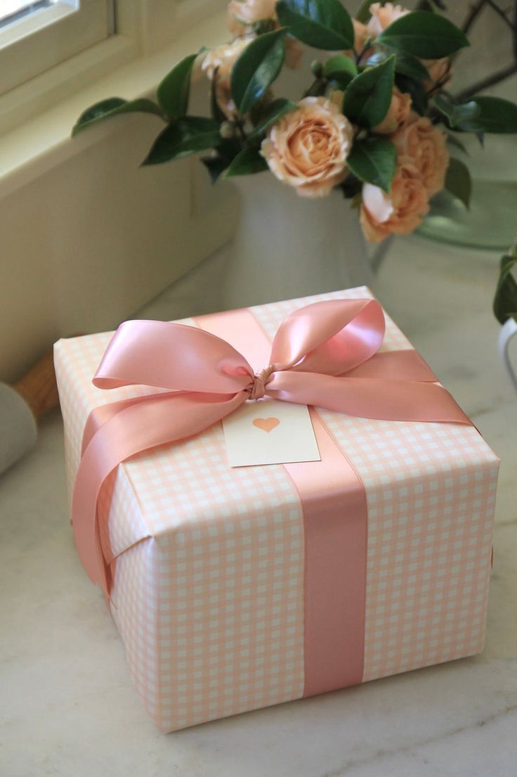 a pink and white checkered gift box with a bow on the top, sitting on a table next to flowers