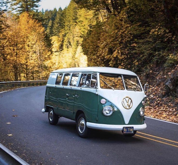 a green and white vw bus driving down a road next to some tall trees