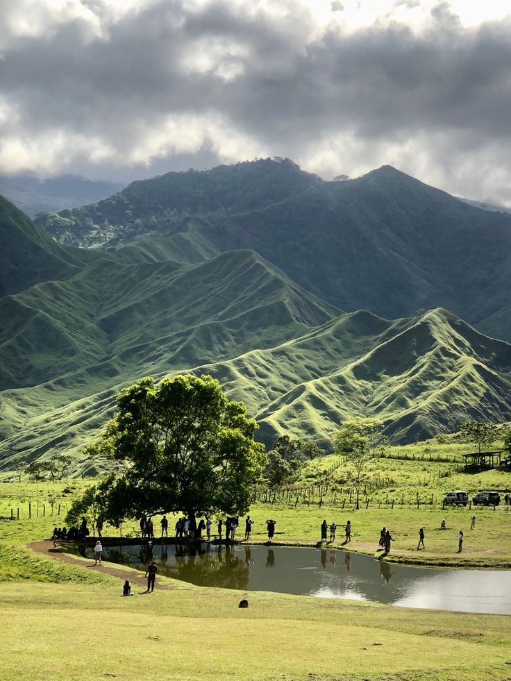 many people are walking around in the grass near a lake and mountains with green hills behind them