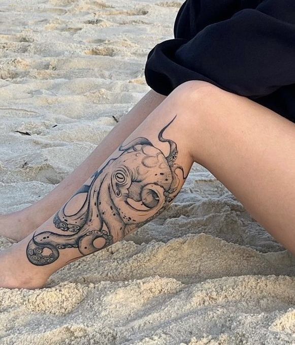 a woman's legs with an octopus tattoo on them sitting in the sand at the beach