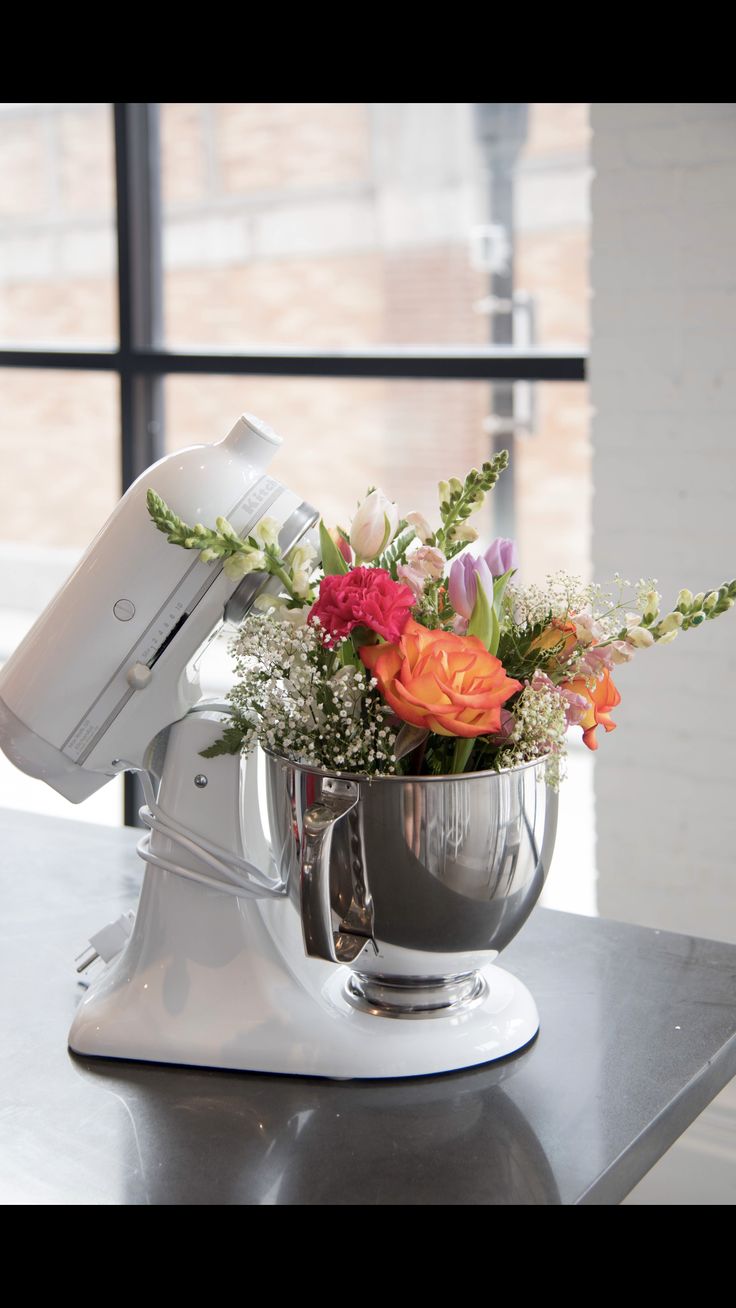 a mixer with flowers in it sitting on a table next to a window and some windows