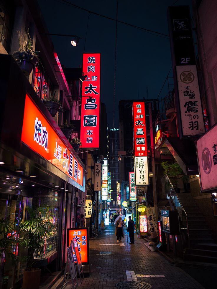 people walking down an alley at night with neon signs on the buildings in the background