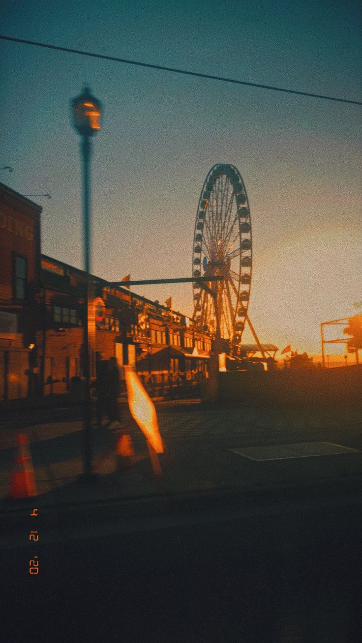 a ferris wheel is in the distance as the sun sets behind it and people are walking by