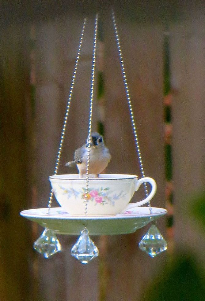 a bird sitting on top of a tea cup hanging from a chain with beads attached to it