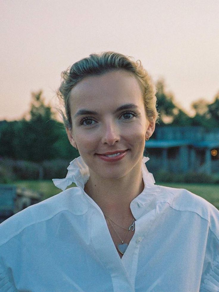 a woman standing in front of a house with trees and grass behind her, smiling at the camera