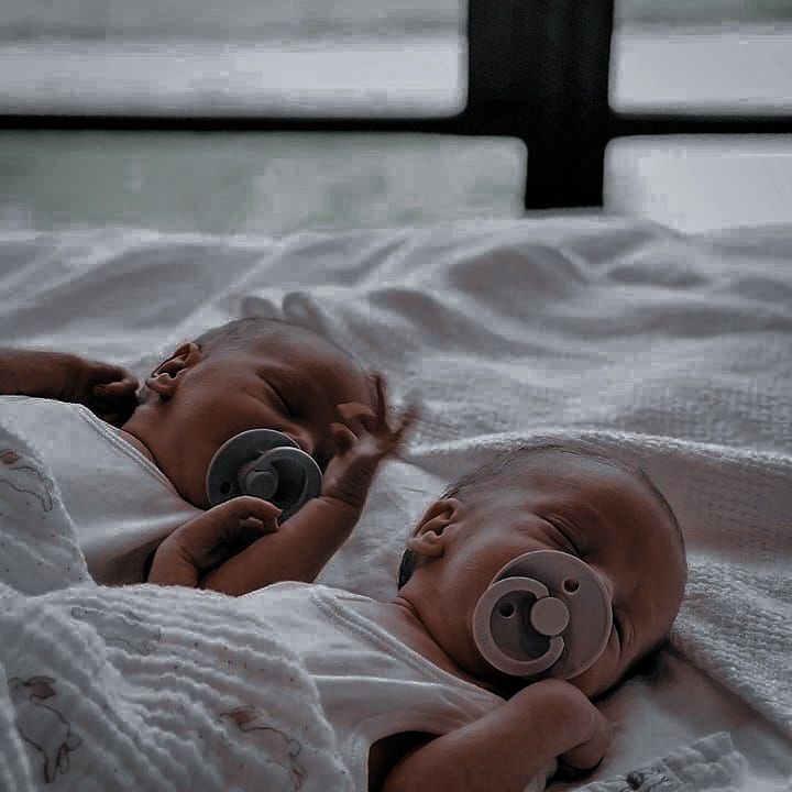 two baby dolls laying on top of a bed next to each other with pacifiers in their mouths