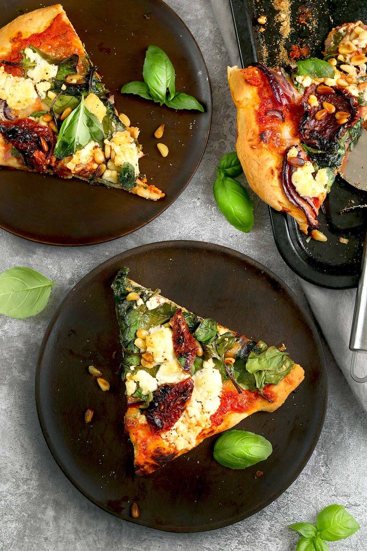 two slices of pizza on black plates with basil and pine nuts next to the plate