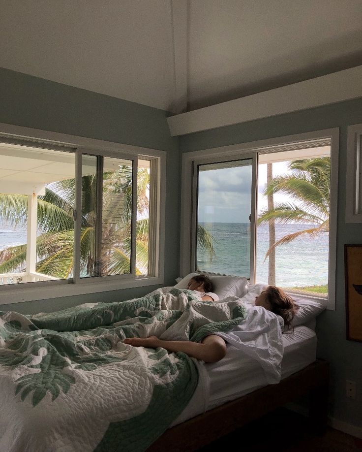 two people laying in bed looking out the window at the ocean and palm trees outside