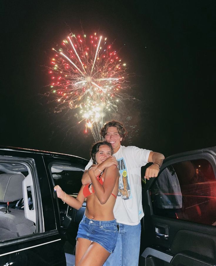 a man and woman standing next to a car with fireworks in the sky behind them