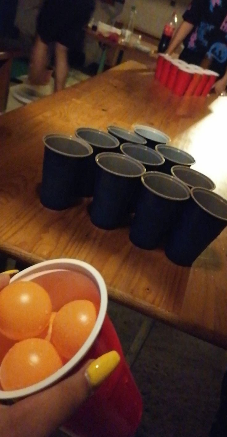 a person holding a cup filled with oranges on top of a wooden table next to cups