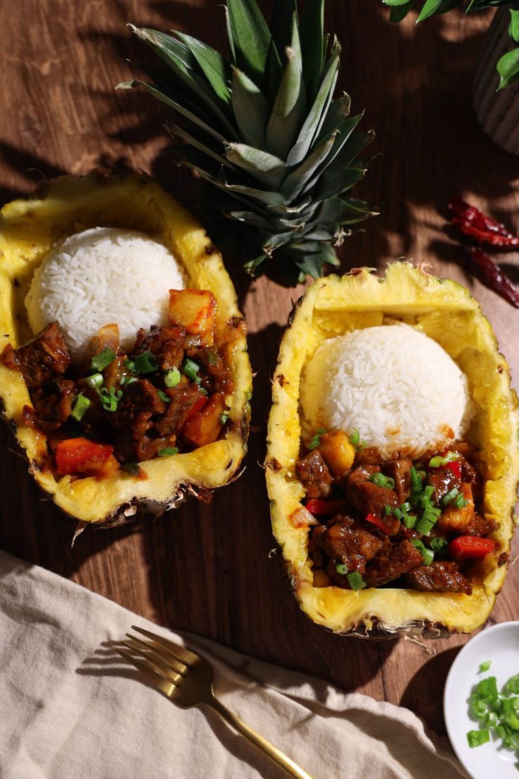 two pineapple halves filled with rice and meat on top of a wooden table next to a fork