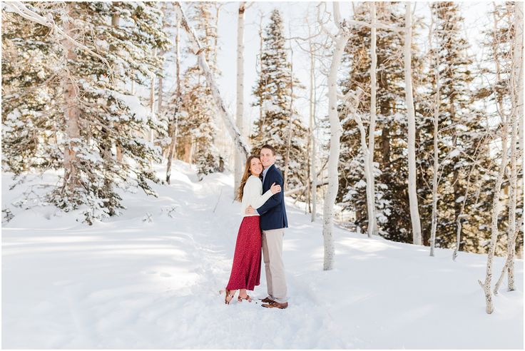 an engaged couple standing in the snow surrounded by trees