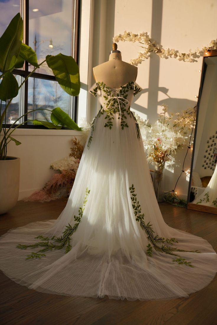 a wedding dress on display in front of a window with flowers and greenery around it