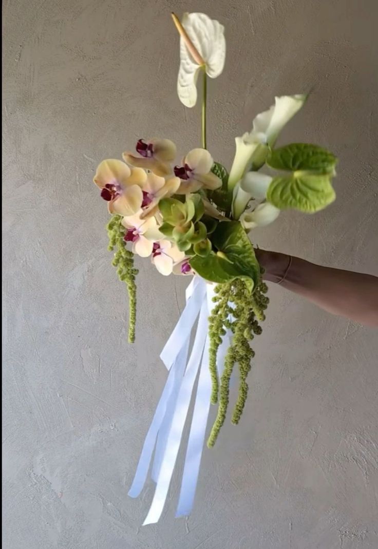 a hand holding a bouquet of flowers with white ribbons and green leaves on the stems