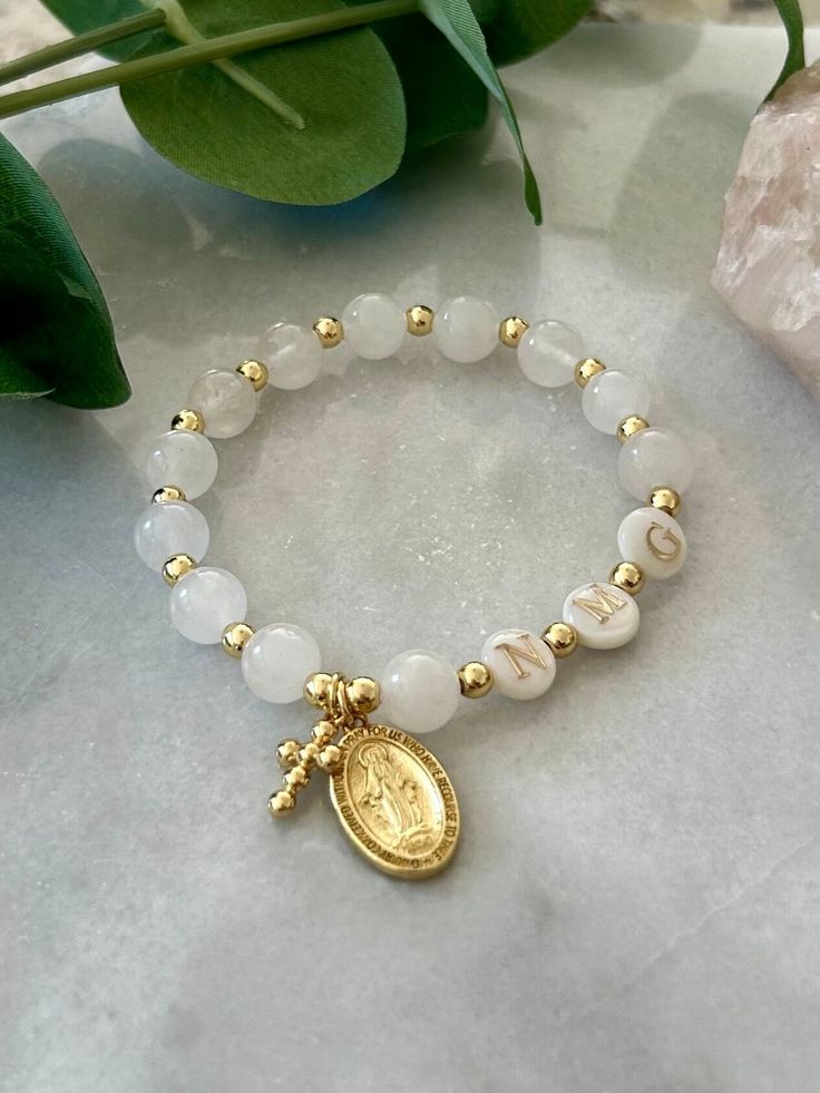 - Beautiful stretch catholic rosary bracelet made with white jade gemstone, pearl initials and 18k gold filled beads. This personalized rosary is adorned with cross charm and Blessed Virgin Mary medal. Bracelet is beaded on high quality stretch cord and secured.  - Rosary bracelet comes in a standard 6.75" size. If you need slightly smaller or larger, please contact me as adjustments may be made. - 8mm white jade beads - 8mm pearl shell initial beads - 4mm gold filled beads - Listing photos incl White Dainty Rosary Bracelet With Round Beads, Dainty White Rosary Bracelet With Round Beads, Personalized White Beaded Bracelets For Baptism, Rosery Bracelet, White Miraculous Medal Spiritual Jewelry, Personalized White Beaded Bracelets For First Communion, Personalized White Spiritual Stretch Bracelet, Adjustable Rosary Bracelet With Round Beads For First Communion, Personalized Spiritual Rosary Bracelet With Round Beads