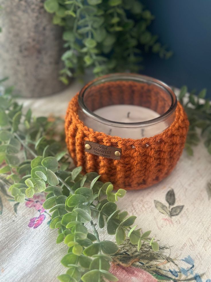 an orange crocheted candle holder sitting on top of a table next to plants