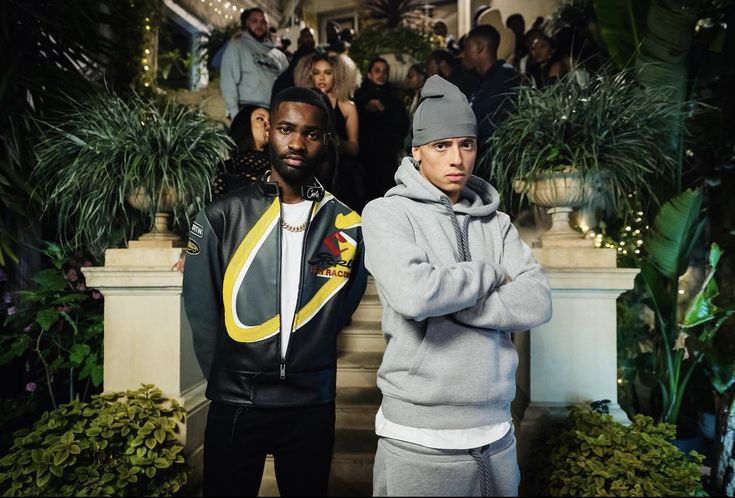 two young men standing next to each other in front of plants and people on stairs