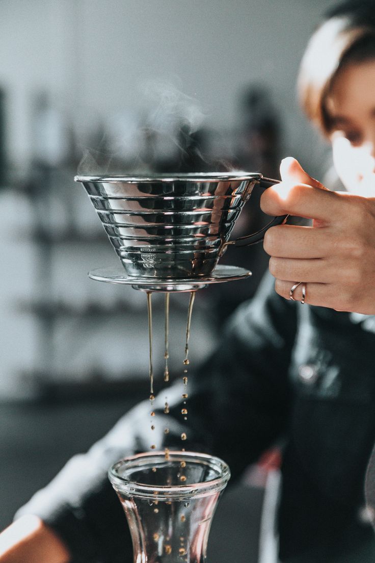 a woman pours coffee into a glass in front of a metal strainer on top of a counter