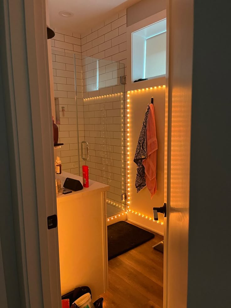 White polished bathroom with glass shower, with soft orange LED lights decorating the edges Cute Bathroom Lighting, Bathroom Mirrors Small Space, Cool Shower Lighting, Bathroom Couple Ideas, Aesthetic Bathroom Shower Ideas, Bedroom Decor College Apartment Inspiration, Bathroom Small Aesthetic, College Apartment Halloween Decor, Mood Lighting For Bathroom