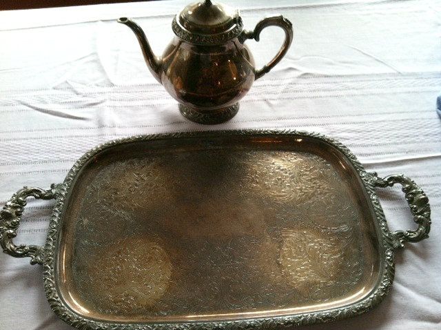 an ornate silver tray with a teapot on the top and white tablecloth behind it