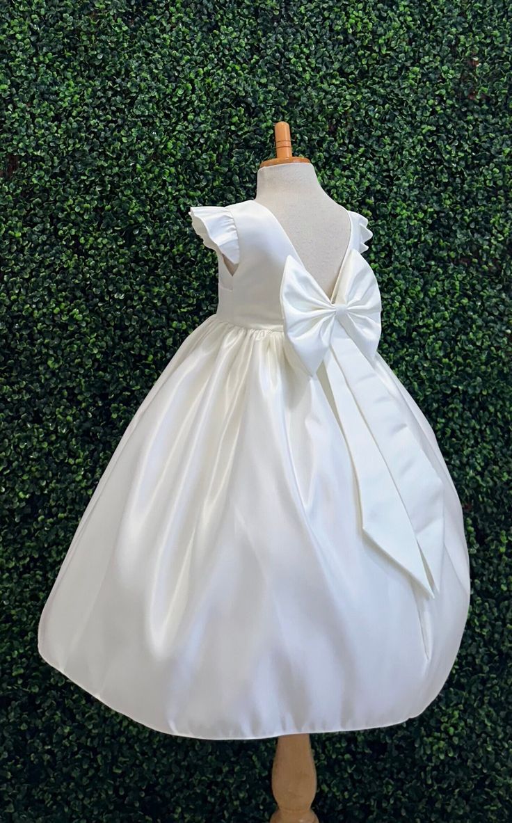 Our enchanting Ankle Length flower girl dresses are sure to turn heads! This beautiful elegant sophisticated dress is handmade, the bodice consists of Ivory Bridal Satin with a sowed in sash at the waist, the back of the dress consists of an open V-back with a hidden zipper. The big bow is detachable. The skirt consists of Ivory satin followed by lining and crinoline for a fuller look. Extremely light weight and beautiful. This dress is perfect for any occasion!  Dress Is Pictured with a pettico Princess-style Baptism Dress With Bow, Elegant First Communion Dress With Ruffles, Elegant Princess Dress With Ruffles For First Communion, Princess Dress With Satin Bow For Baptism, Elegant Fitted Princess Dress For Baptism, Elegant Cream Princess Dress For Wedding, Elegant Baptism Dress With Bow For Party, Cream Princess Dress For First Communion, Fitted Cream Princess Dress For First Communion