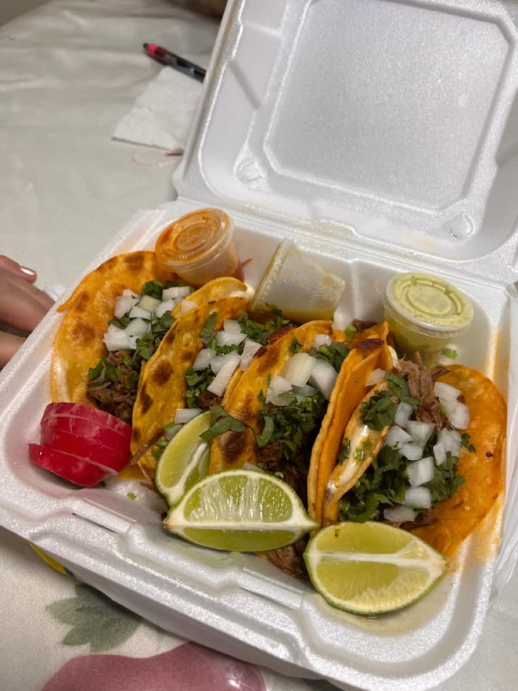 three tacos in a foam container with limes, onions and cilantro