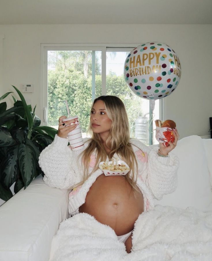 a pregnant woman sitting on top of a white couch eating food and drinking from cups