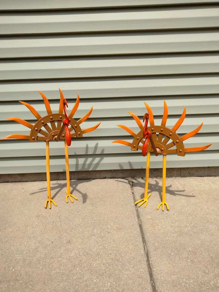 two metal birds are standing on legs with orange tails and feet, one is facing the other