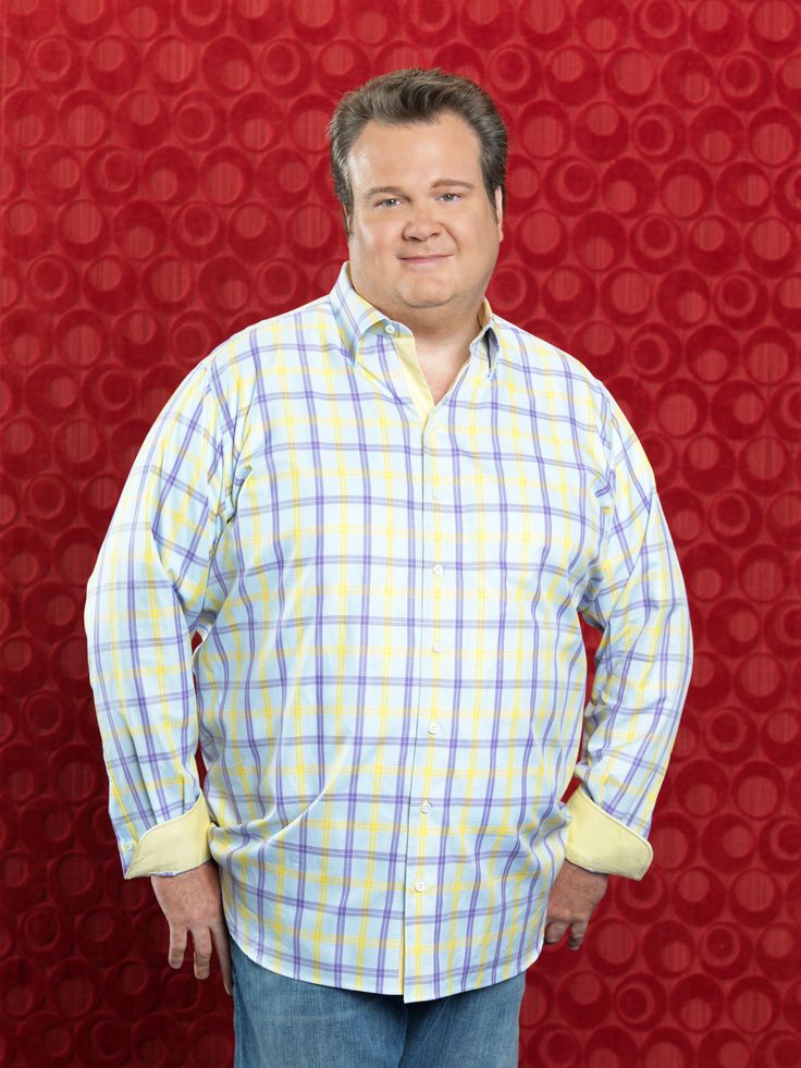 a man standing in front of a red wall wearing a plaid shirt and blue jeans