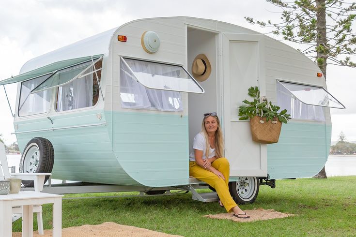 a woman sitting in the doorway of an old camper trailer with potted plants