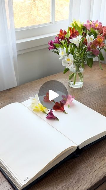 an open book sitting on top of a wooden table next to a vase with flowers