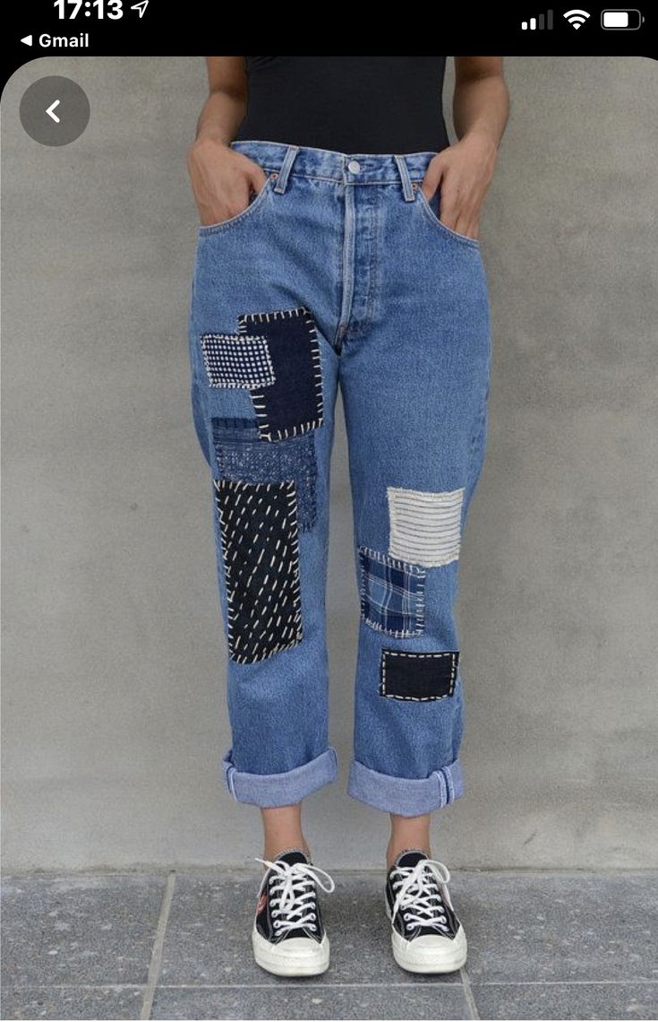 a woman standing in front of a wall wearing blue jeans with patches on the side