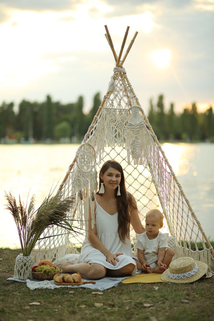 a woman and her baby sitting in front of a teepee next to the water