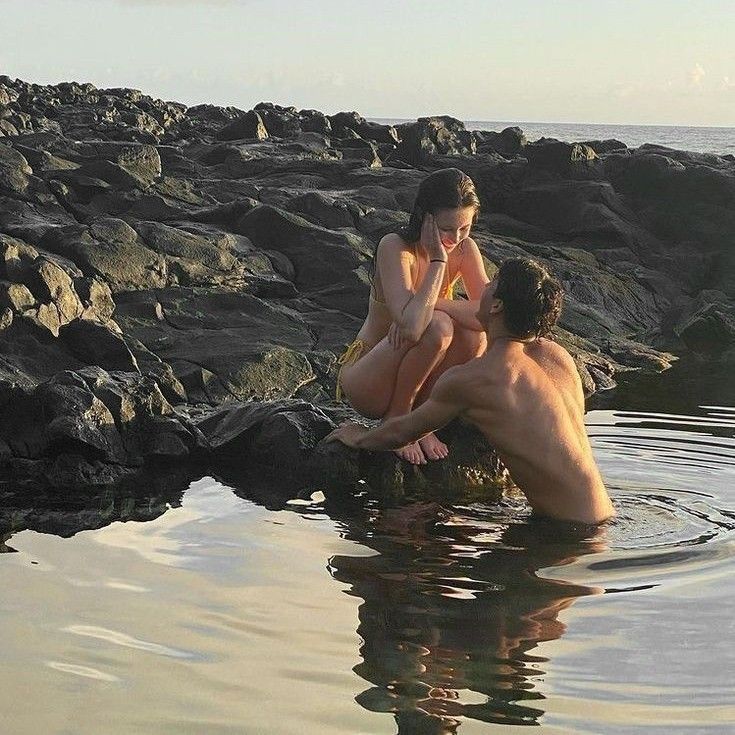 two people are sitting in the water near some rocks and one is talking on a cell phone