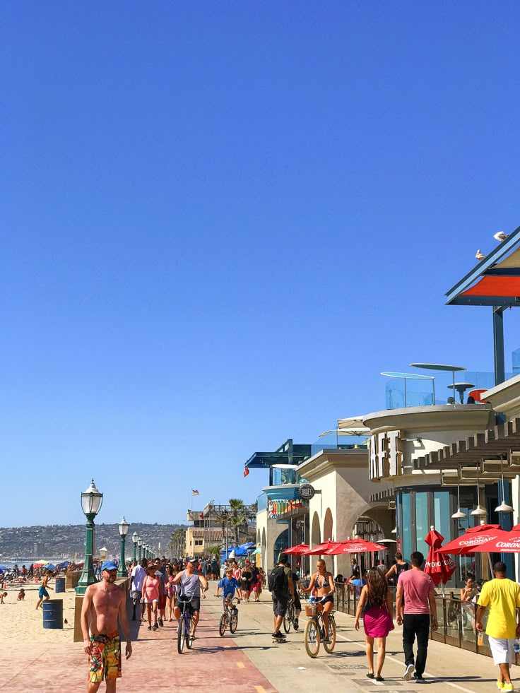 many people are walking and riding bikes on the sidewalk near the beach with umbrellas