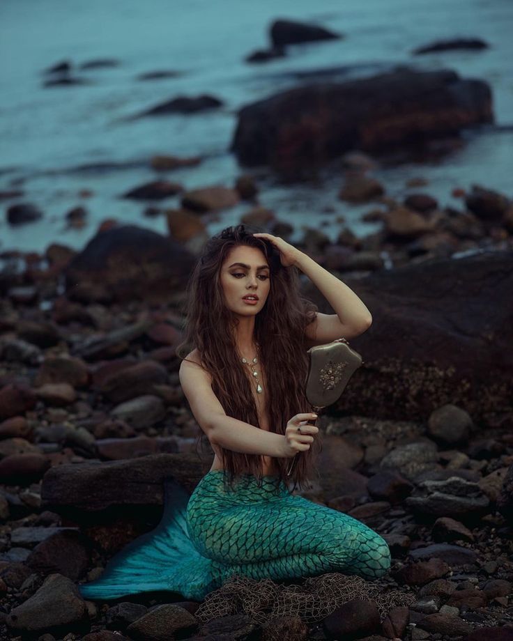 a woman with long hair sitting on rocks next to the ocean and holding a book