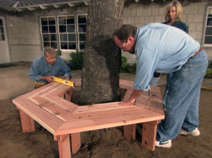 two people working on a wooden bench in front of a tree and a woman standing next to it